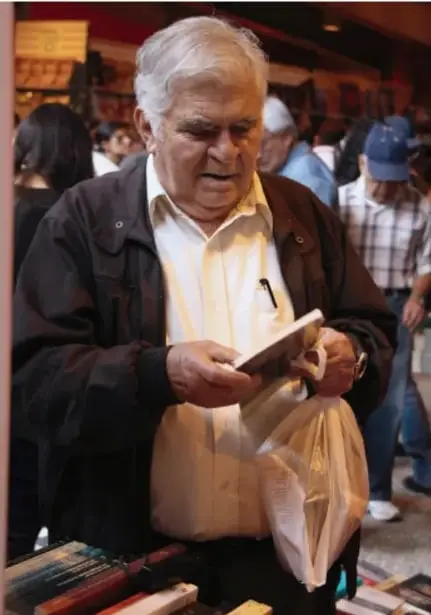 Fotografía de un hombre mayor hojeando un libro en un puesto de venta al aire libre, con otras personas alrededor.