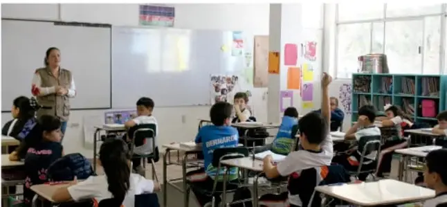 Aula de primaria con una maestra frente al pizarrón y estudiantes trabajando en sus pupitres.