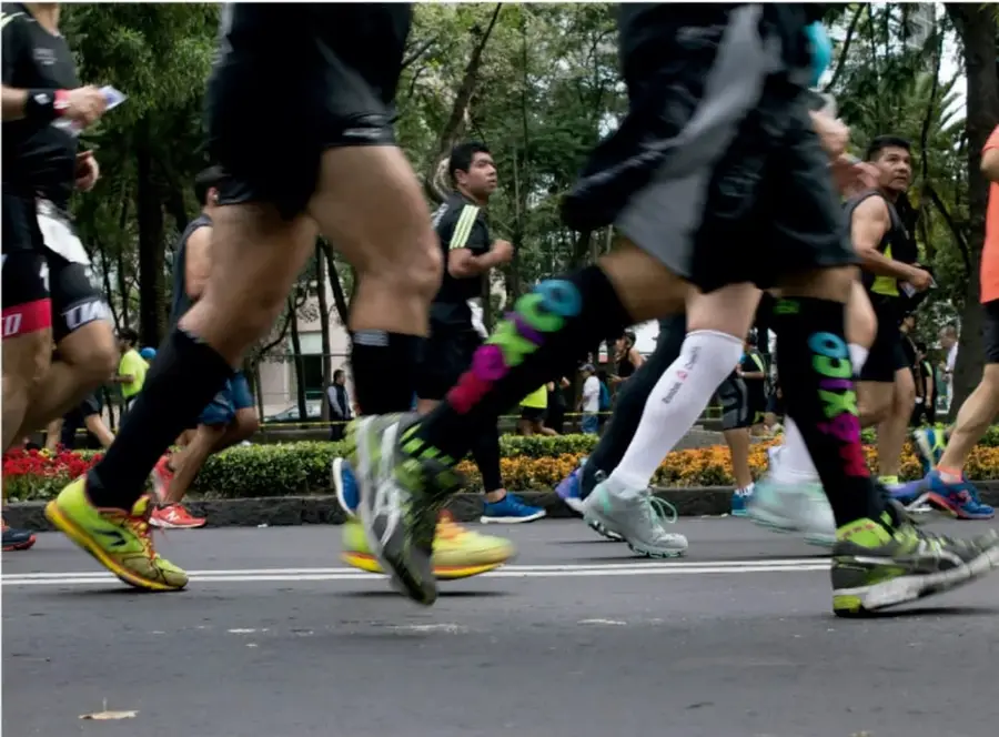 Fotografía de corredores en una carrera urbana: se ven piernas en movimiento, zapatillas de colores, medias de compresión y un parque con árboles y flores al fondo, ilustrando la ejecución de una actividad grupal.