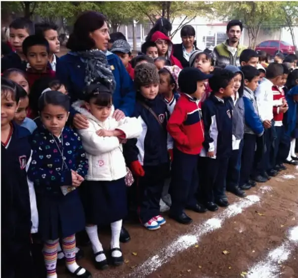 Niñas y niños de primaria formados en una cancha, con abrigos y uniformes, acompañados por una maestra.
