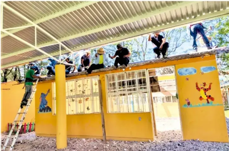 Fotografía de miembros de una comunidad trabajando en la construcción/reparación de un edificio comunitario amarillo con techo metálico, algunos en escaleras y otros sobre la estructura.