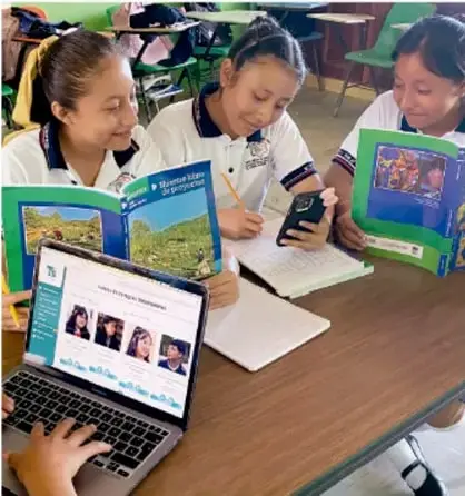 Fotografía de tres estudiantes de telesecundaria trabajando en equipo alrededor de una mesa con libros, cuadernos y una laptop, consultando información para el proyecto.