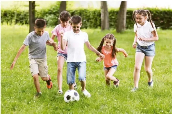 Fotografía de cinco niños y niñas jugando fútbol en un parque, corriendo detrás de un balón sobre el césped.