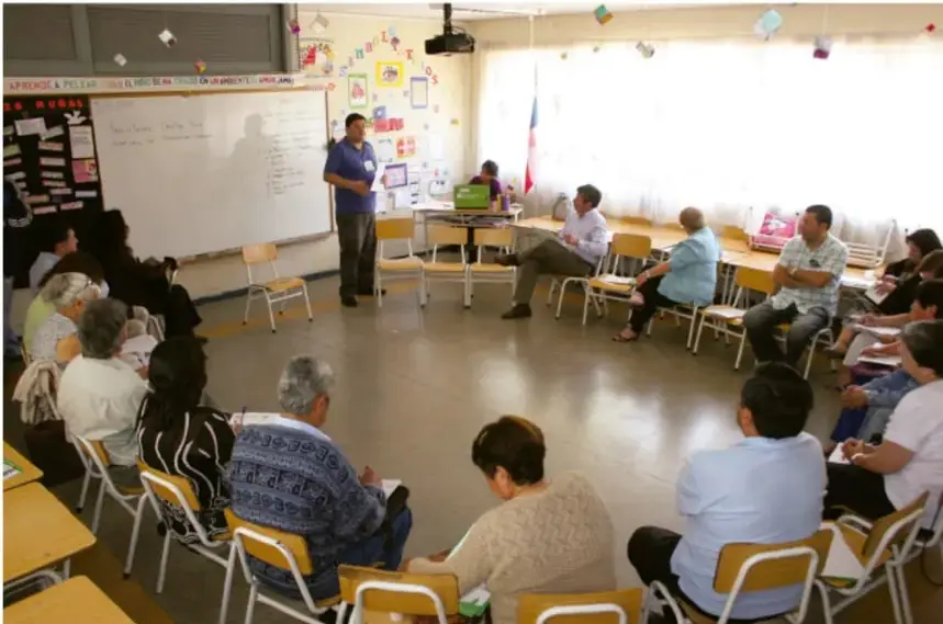 Fotografía de un aula con un grupo de personas sentadas en círculo participando en una sesión de interacción y aprendizaje, con un facilitador de pie frente a un pizarrón.
