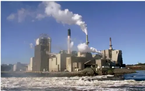 Fotografía de una planta industrial con chimeneas que emiten humo, ubicada junto a un cuerpo de agua, ilustrando los procesos productivos y su impacto ambiental.