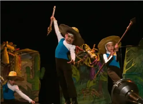 Fotografía de tres niños actores sobre un escenario: llevan sombreros de charro, chalecos azules y bastones; al fondo hay escenografía con cactus y un caballo, representando una obra teatral.