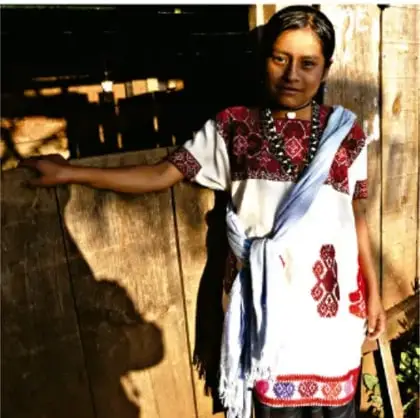 Fotografía de una mujer indígena con blusa bordada y rebozo, apoyada en una pared de madera.