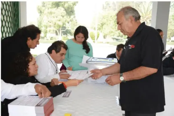 Reunión de personas alrededor de una mesa con documentos; un hombre de pie dirige la actividad, en un espacio con ventanales.