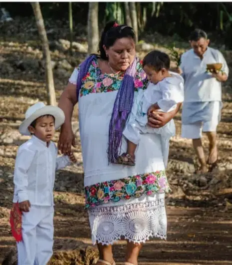 Fotografía de una comunidad maya: una madre viste traje blanco tradicional bordado y carga a un niño pequeño; otro niño con sombrero camina al lado; al fondo un hombre mayor con ropa blanca porta un recipiente, posiblemente parte de la ceremonia.