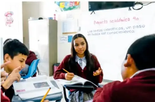 Fotografía de estudiantes en el aula: una chica explica frente a sus compañeros junto a un rotafolio con el título 'Proyecto académico 4.0: Los temas científicos, un paso en busca del conocimiento'.
