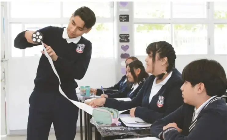 Fotografía de un aula de telesecundaria: una estudiante de uniforme se para frente a sus compañeros mostrando una tira larga de papel con registro escrito, mientras cuatro compañeros sentados en sus mesas observan, escuchan y toman notas.