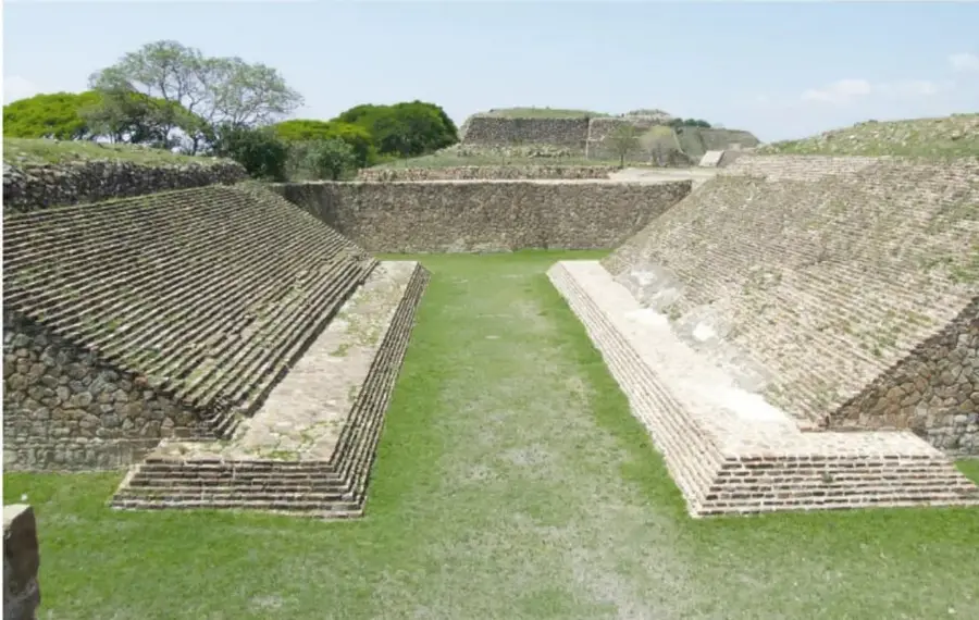 Fotografía de una cancha de juego de pelota arqueológica: explanada larga de pasto entre dos muros inclinados de piedra, con edificios y árboles al fondo.