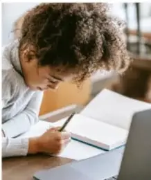 Estudiante escribiendo en un cuaderno frente a una laptop, con expresión concentrada.