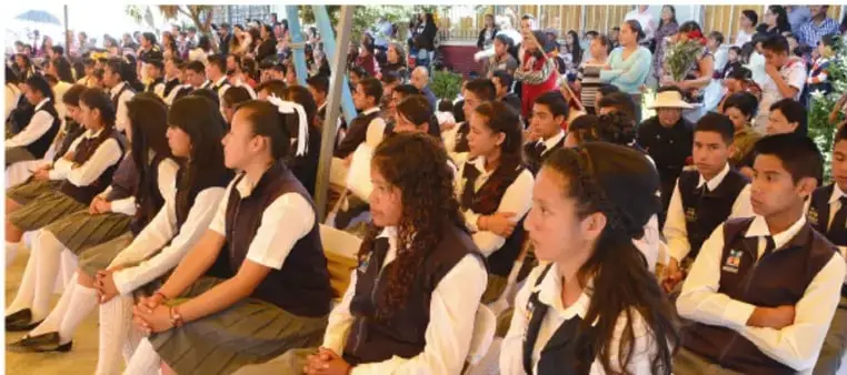 Fotografía de estudiantes de telesecundaria uniformados, sentados en gradas o bancas en un acto escolar, atentos y participando en una actividad colectiva.