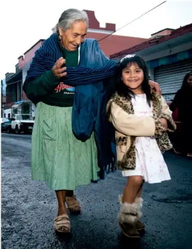 Fotografía de una adulta mayor y una niña caminando juntas por una calle, de brazos, en actitud de cercanía y acompañamiento.