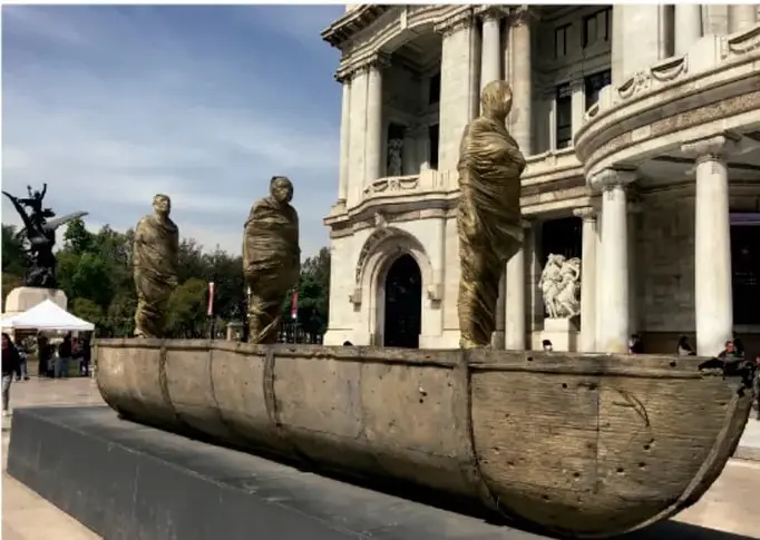 Fotografía de la escultura en bronce El ruido generado por el choque de los cuerpos, de Jorge Marín: una barca de madera con tres figuras humanas cubiertas de tela, ubicada frente a un edificio con columnas.