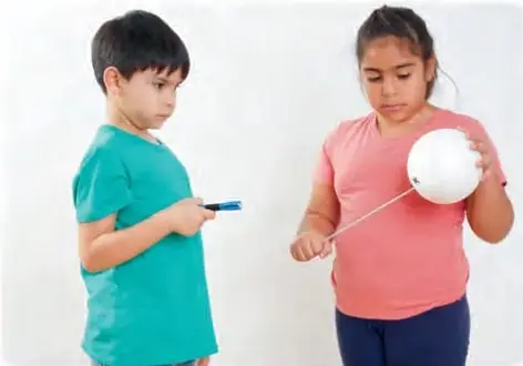 Niño y niña manipulando una pelota de plumavit con un palillo enterrado para hacerla girar; icono de precaución 'Manipulen el palillo con mucho cuidado'.