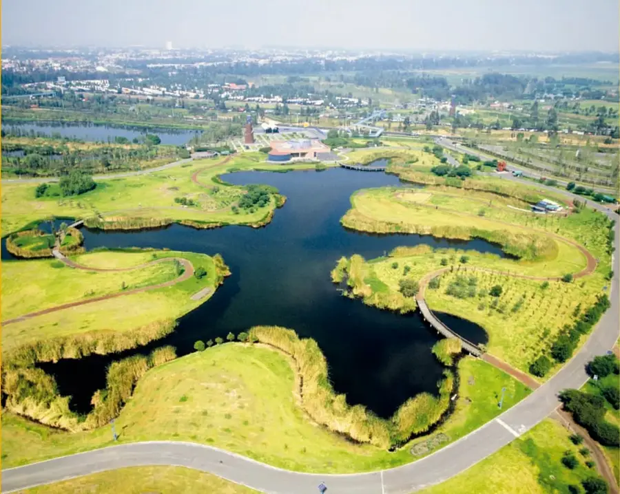 Fotografía aérea de un parque urbano con lagos, jardines con senderos sinuosos y caminos empedrados, rodeado de vegetación y zonas residenciales al fondo.