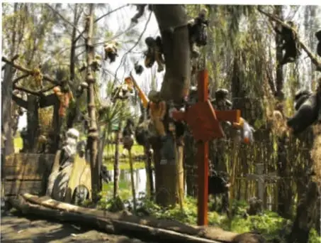 Fotografía de un paraje natural con árboles, una cruz de madera roja y ofrendas colgadas; al fondo más cruces y ramas.