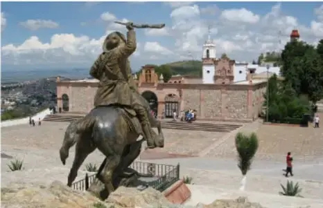 Monumento ecuestre de Francisco 'Pancho' Villa a caballo con el brazo en alto, ubicado en la Plaza de Armas de la ciudad de Chihuahua, con edificios coloniales al fondo.