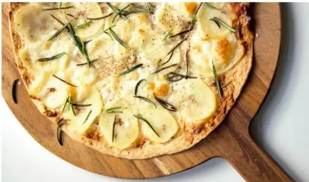 Fotografía de una masa o focaccia horneada decorada con rodajas de papa y hierbas, servida sobre una tabla de madera, ilustrando la papa como ingrediente común.