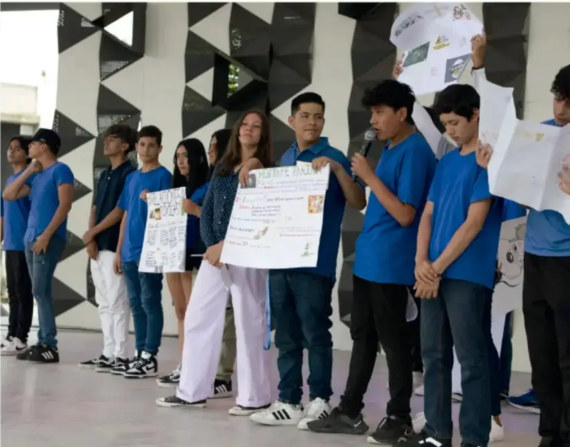 Fotografía de un grupo de estudiantes de secundaria con camisetas azules que presentan carteles con los resultados de su proyecto frente a la comunidad escolar; uno de ellos habla con un micrófono.