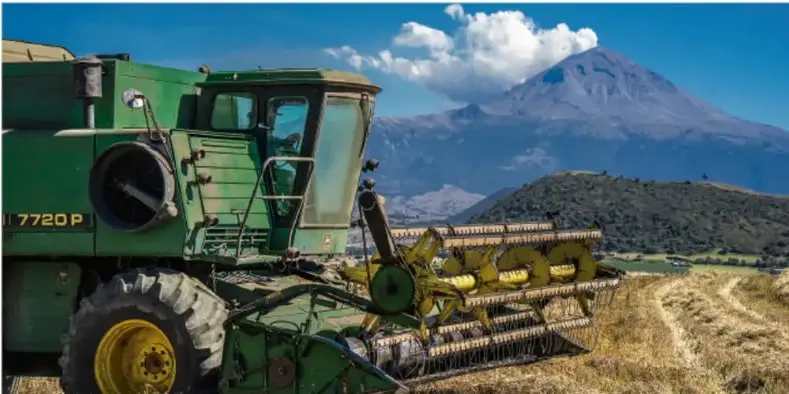Cosechadora verde (modelo 7720P) trabajando en un campo con un volcán nevado al fondo.