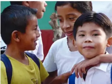 Fotografía de tres niños sonrientes, uno con playera amarilla y otro con playera blanca, en un ambiente escolar.