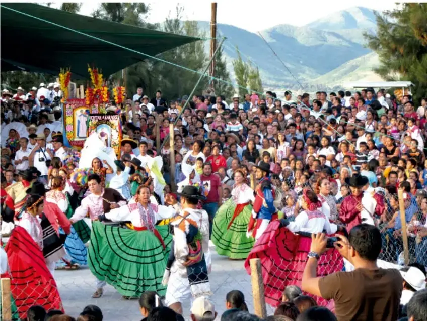 Fotografía de una fiesta o celebración comunitaria en una localidad andina mexicana: mujeres con faldas de colores verdes, rojas y azules bailan en un espacio abierto, rodeadas de una multitud de asistentes, con montañas y una carpa verde al fondo; se observa un aparato o estandarte religioso decorado.