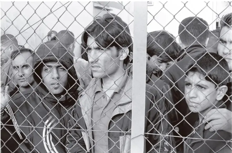 Fotografía en blanco y negro de un grupo de personas, entre ellas niños y adultos, detrás de una malla ciclónica; miran hacia el frente con expresión seria.