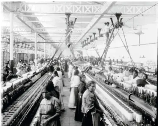 Fotografía en blanco y negro de una fábrica textil durante el Porfiriato. Se observan hileras de máquinas y trabajadores, en su mayoría mujeres, operando los telares en un amplio galpón industrial.