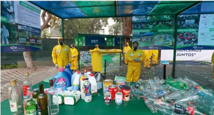 Foto de una estación de reciclaje al aire libre con personas amarillas de protección, contenedores PET, botellas plásticas, envases de vidrio y carteles sobre reciclaje y aprovechamiento de residuos.
