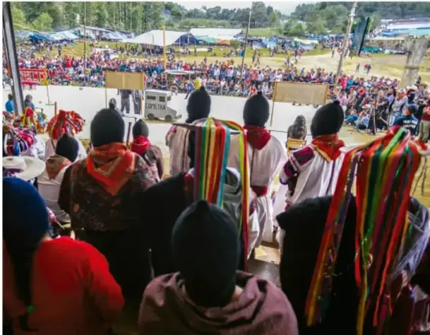 Fotografía de una asamblea comunitaria al aire libre con numerosos asistentes, muchos con vestimenta tradicional y sombreros, reunidos en un espacio público.
