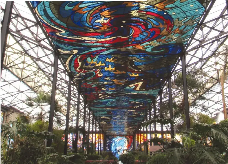 Vitrales de gran formato con figuras geométricas y orgánicas en tonos azules, rojos, amarillos y naranjas, montados sobre una estructura metálica que cubre un pasillo con vegetación.