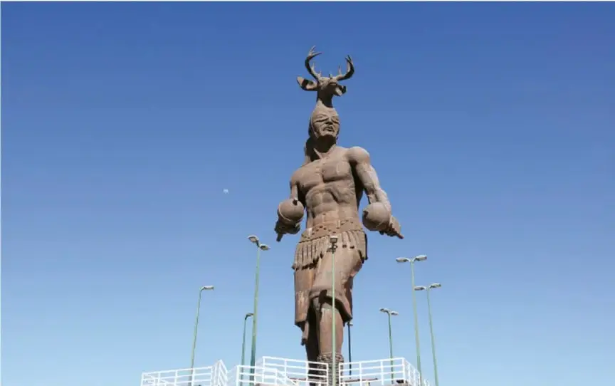 Fotografía de una estatua monumental de un guerrero indígena con penacho de venado, sobre una plataforma, bajo cielo azul con farolas alrededor (monumento a Cuauhtémoc o figura similar).