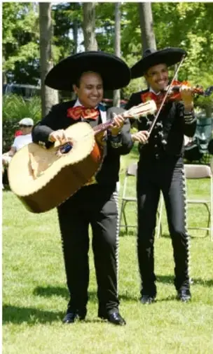 Fotografía de dos músicos de mariachi con traje charro y sombrero, tocando guitarrón y violín al aire libre, en un jardín con árboles al fondo.