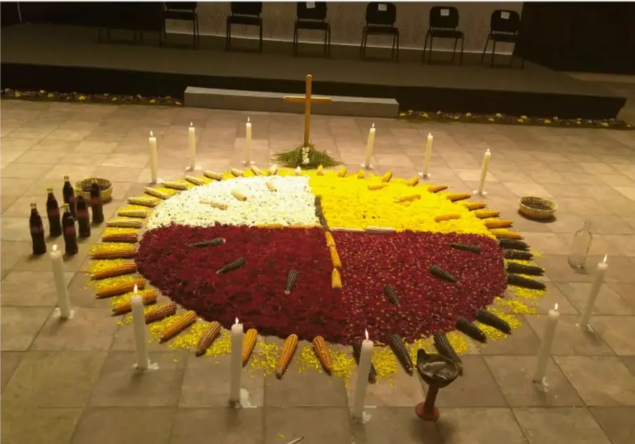 Fotografía de un altar circular de los mayas tseltales: un gran círculo en el piso dividido en cuatro cuadrantes de pétalos de colores (blanco, amarillo, rojo y verde oscuro), con mazorcas dispuestas radialmente en el borde, una cruz de madera con un manojo de plantas en el centro, velas encendidas alrededor y ofrendas (botellas, incensario) sobre un piso de piedra, en un espacio techado.