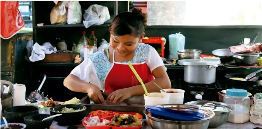 Fotografía de una cocinera con delantal rojo preparando alimentos en una cocina tradicional rodeada de ollas, ingredientes y utensilios; ilustra la preparación de comida como práctica cultural y artesanal.