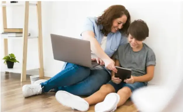 Fotografía de una madre y su hijo sentados en el piso usando una laptop y una tableta, como ejemplo de comunicación a distancia.