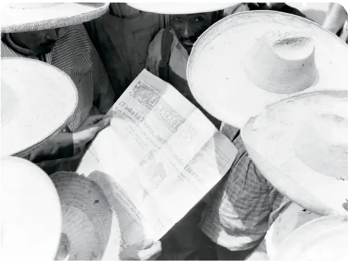 Fotografía en blanco y negro de varios campesinos con grandes sombreros de palma reunidos alrededor de un periódico que uno sostiene; pie: 'Campesinos leyendo El Machete, 1929'.