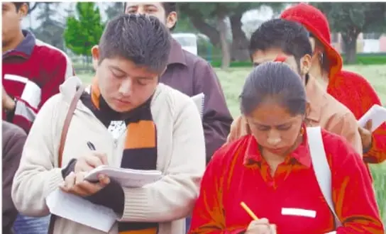 Fotografía de un grupo de estudiantes en exterior, con uniformes rojos y azules, tomando notas en cuadernos; dos de ellos escriben con lápiz y pluma.