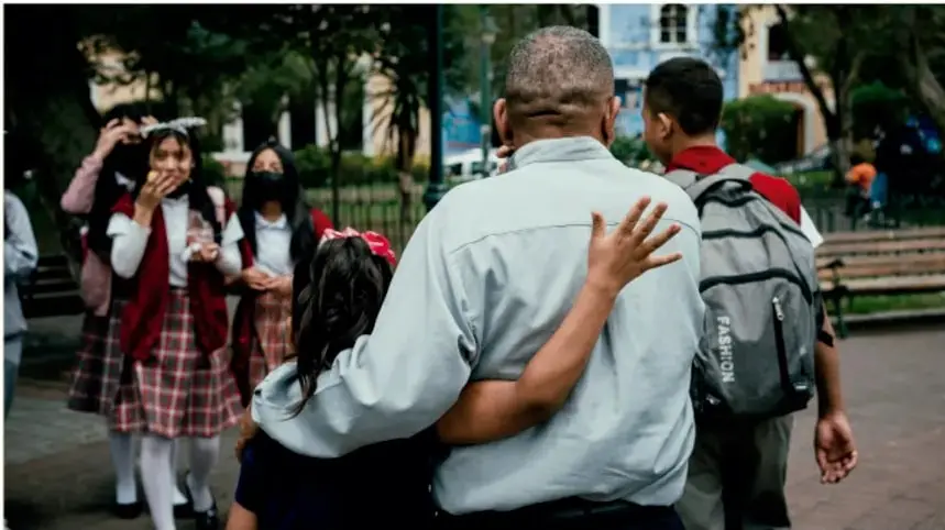 Fotografía de una persona mayor abrazando a una niña en un espacio público, con estudiantes de uniforme al fondo, representando la convivencia y la inclusión social.