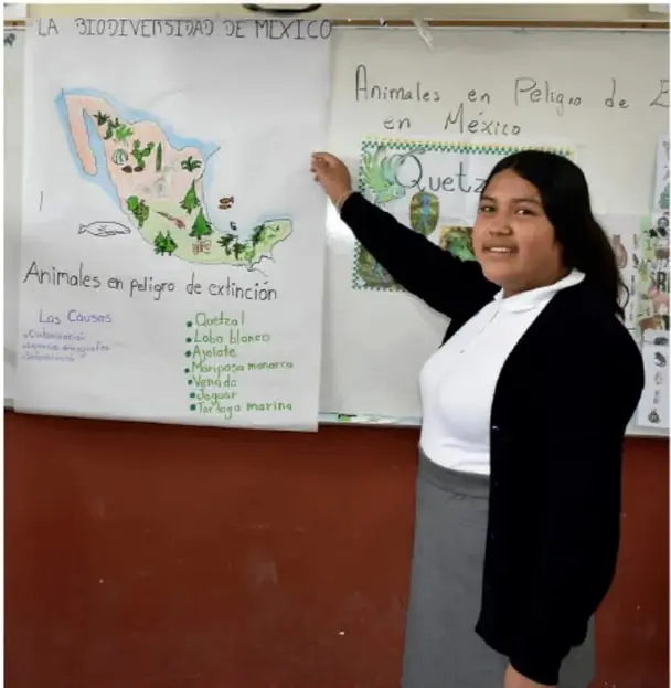 Fotografía de una estudiante de Telesecundaria sonriendo y señalando un periódico mural escolar. El mural lleva el título «La biodiversidad de México» con un mapa del país y un recuadro «Animales en peligro de extinción en México» que enumera: Quetzal, Lobo blanco, Axolote, Monapa monoaca, Venado y Tortuga marina, junto con las causas de su peligro. Crédito: Arturo Morales Rangel, CIMAVEMTE.