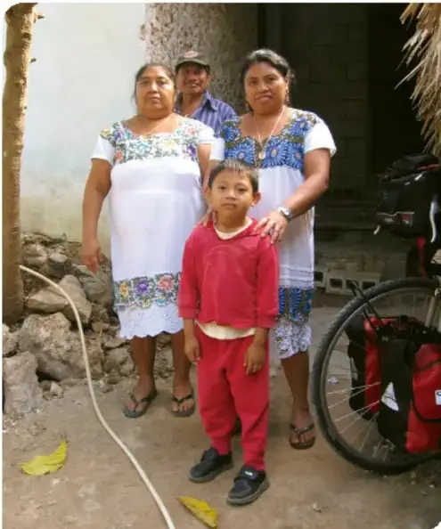 Fotografía de una familia mexicana (dos mujeres con huipiles blancos, un hombre y un niño con suéter rojo) junto a una bicicleta cargada, en un patio con tierra y pared de adobe.
