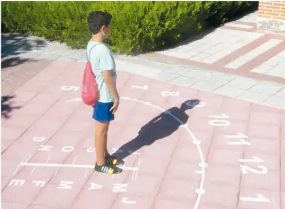 Fotografía de un niño con mochila rosa parado sobre un reloj de sol humano pintado en el piso rosa de un patio: se ven números (6, 7, 8, 10, 11, 12) y nombres de meses pintados; la sombra del niño funciona como manecilla.