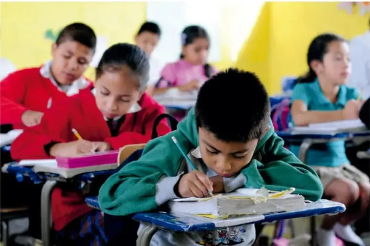 Fotografía de estudiantes de primaria, con uniforme escolar, escribiendo en sus cuadernos dentro de un salón de clases con paredes amarillas.