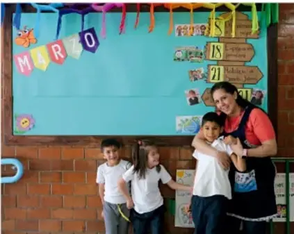 Fotografía de una maestra abrazando a un niño frente a un mural escolar con la palabra MUR y números; otras dos niñas están al lado. Representa a la autoridad escolar cuidando al alumnado.