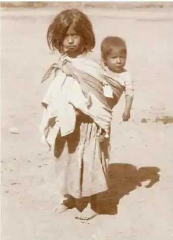Fotografía en blanco y negro de una niña indígena cargando a un bebé en la espalda con un rebozo, de pie sobre un terreno árido. Ilustra la diversidad cultural y las condiciones de vida de la niñez en comunidades originarias.