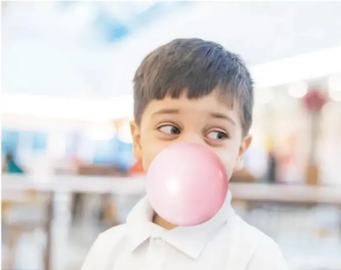 Niño de camisa blanca inflando un globo rosa de chicle, con un fondo difuminado.