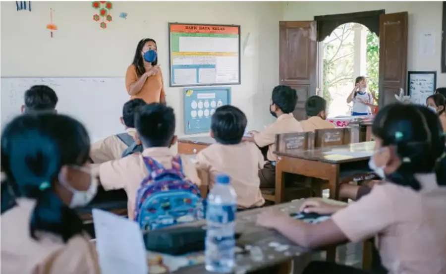 Fotografía de un salón de clases: una maestra de pie con cubrebocas habla frente al grupo, estudiantes sentados en bancas con cubrebocas escuchan; al fondo, un pizarrón con un cartel y un estudiante de pie junto a la puerta.